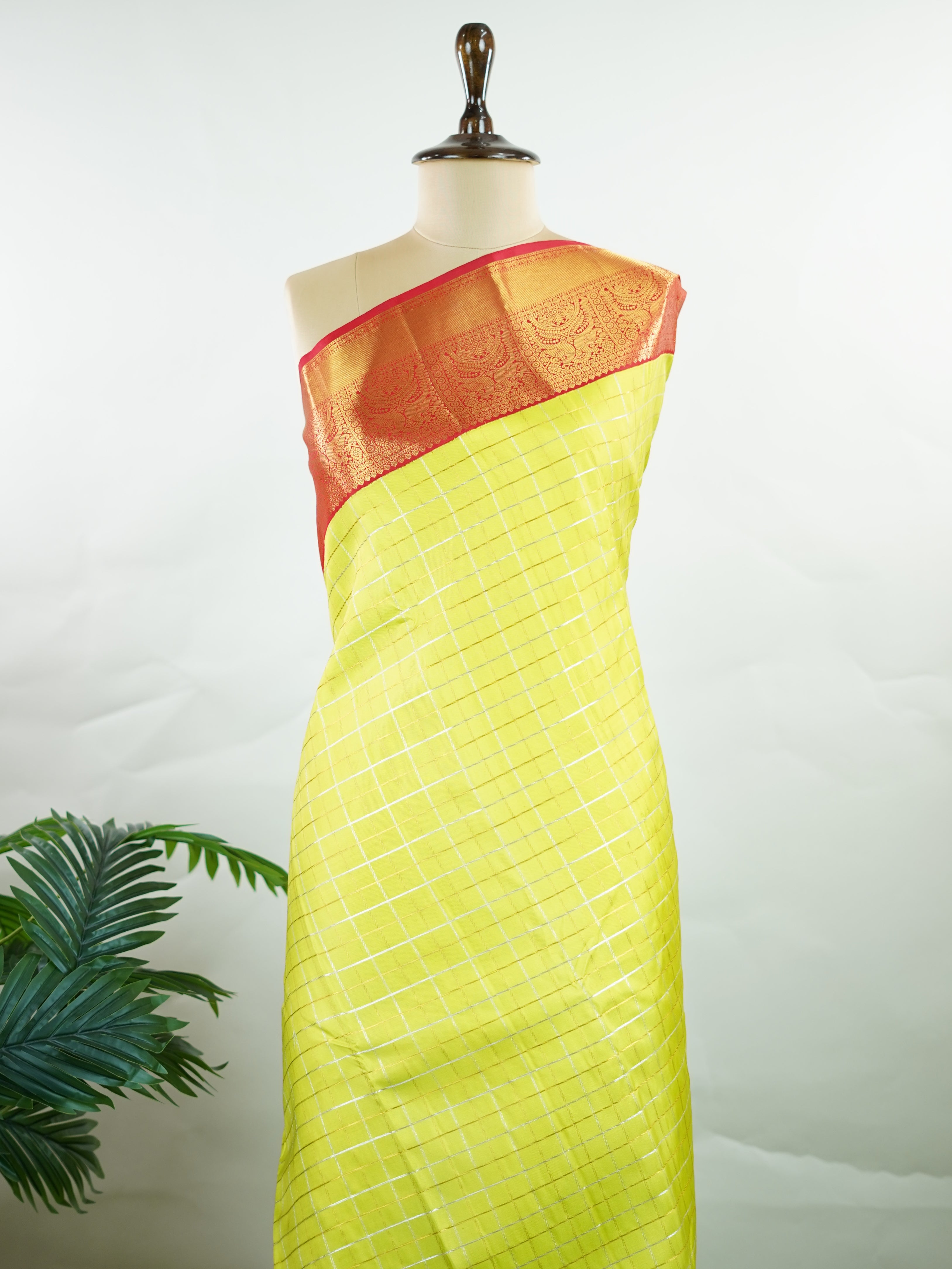 Kanchipuram Silk Beautiful traditional Kanjivaram saree featuring a vibrant Leaf Green body adorned with exquisite gold and silver checks, creating a rich and radiant appearance. The striking Red border is embellished with majestic Annapakshi motifs, adding a touch of heritage and timeless elegance. An intricately woven pallu with classic diamond motifs enhances the grandeur and sophistication of the drape. Paired with a plain Red blouse, this masterpiece is perfect for weddings, festive occasions, and trad