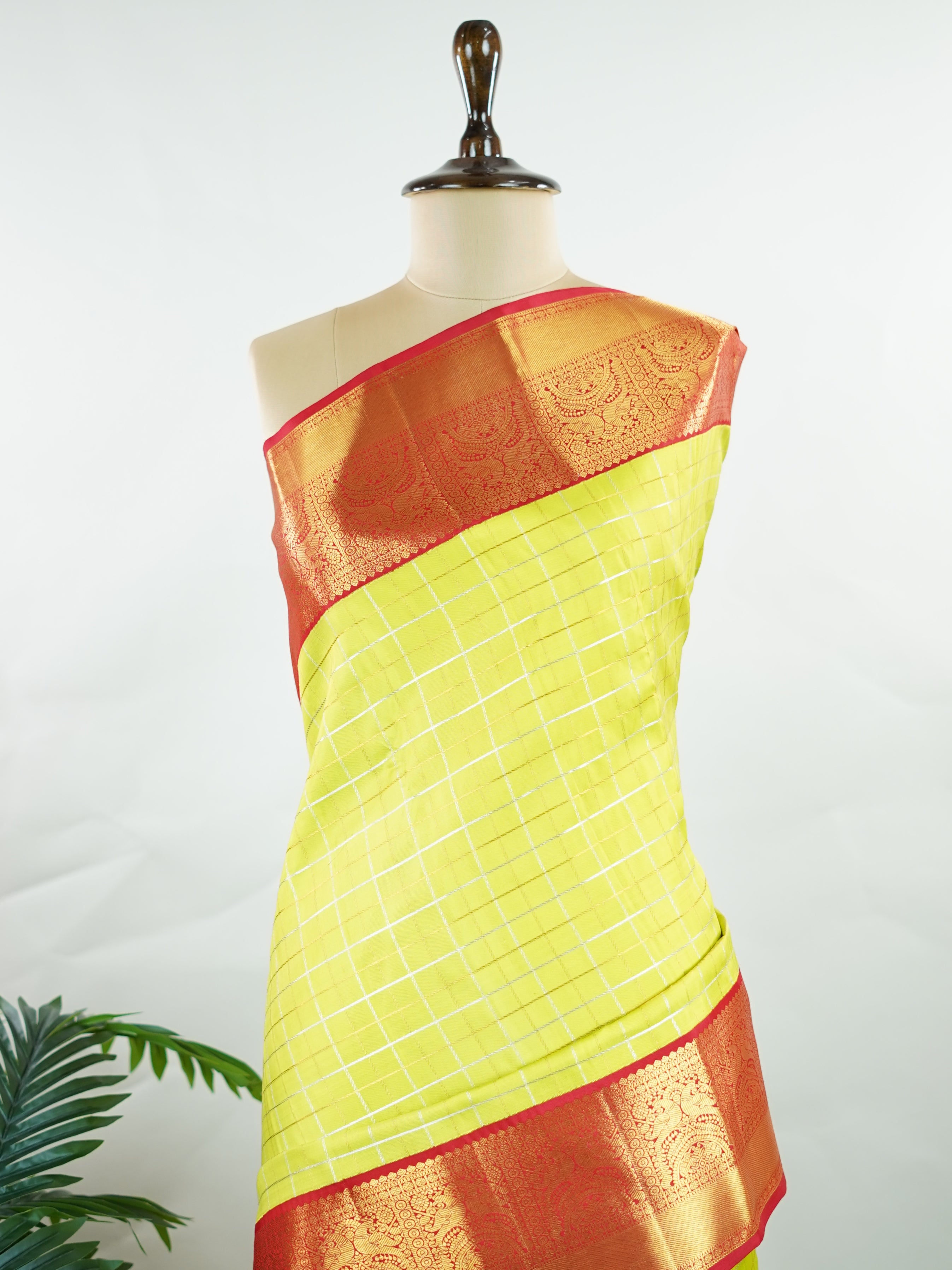 Kanchipuram Silk Beautiful traditional Kanjivaram saree featuring a vibrant Leaf Green body adorned with exquisite gold and silver checks, creating a rich and radiant appearance. The striking Red border is embellished with majestic Annapakshi motifs, adding a touch of heritage and timeless elegance. An intricately woven pallu with classic diamond motifs enhances the grandeur and sophistication of the drape. Paired with a plain Red blouse, this masterpiece is perfect for weddings, festive occasions, and trad