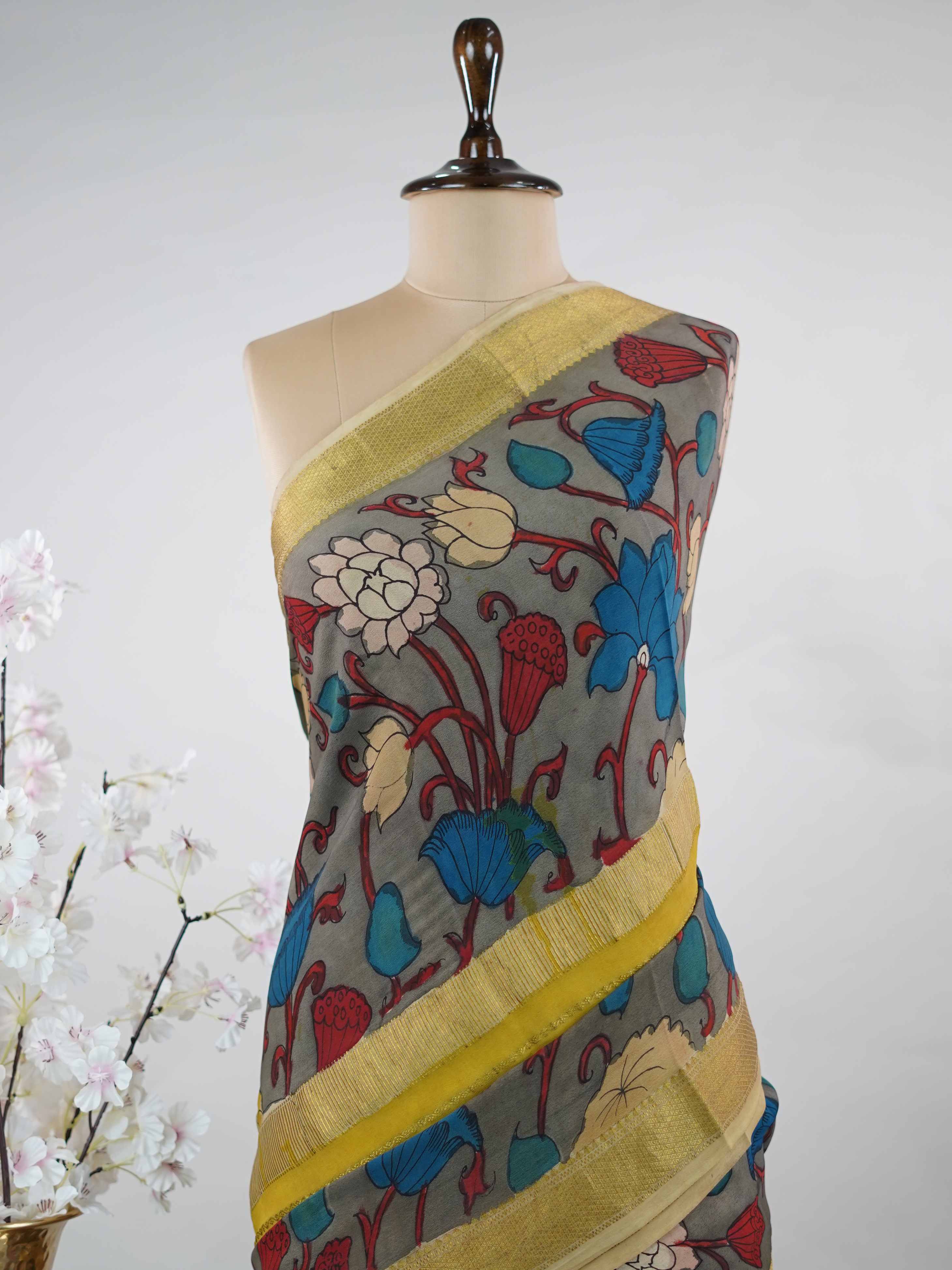 Mysore silk This Mysore silk saree is a quiet celebration of two worlds beautifully united — one half draped in the cool, understated elegance of pure grey silk, the other brought alive with intricate pen Kalamkari artistry in earthy, hand-drawn motifs. The 100% pure silk body carries that characteristic Mysore sheen, smooth and weightless against the skin, while the Kalamkari half tells a story in every careful line and curve. Zari accents frame the drape with a subtle gleam, never overpowering the artistr