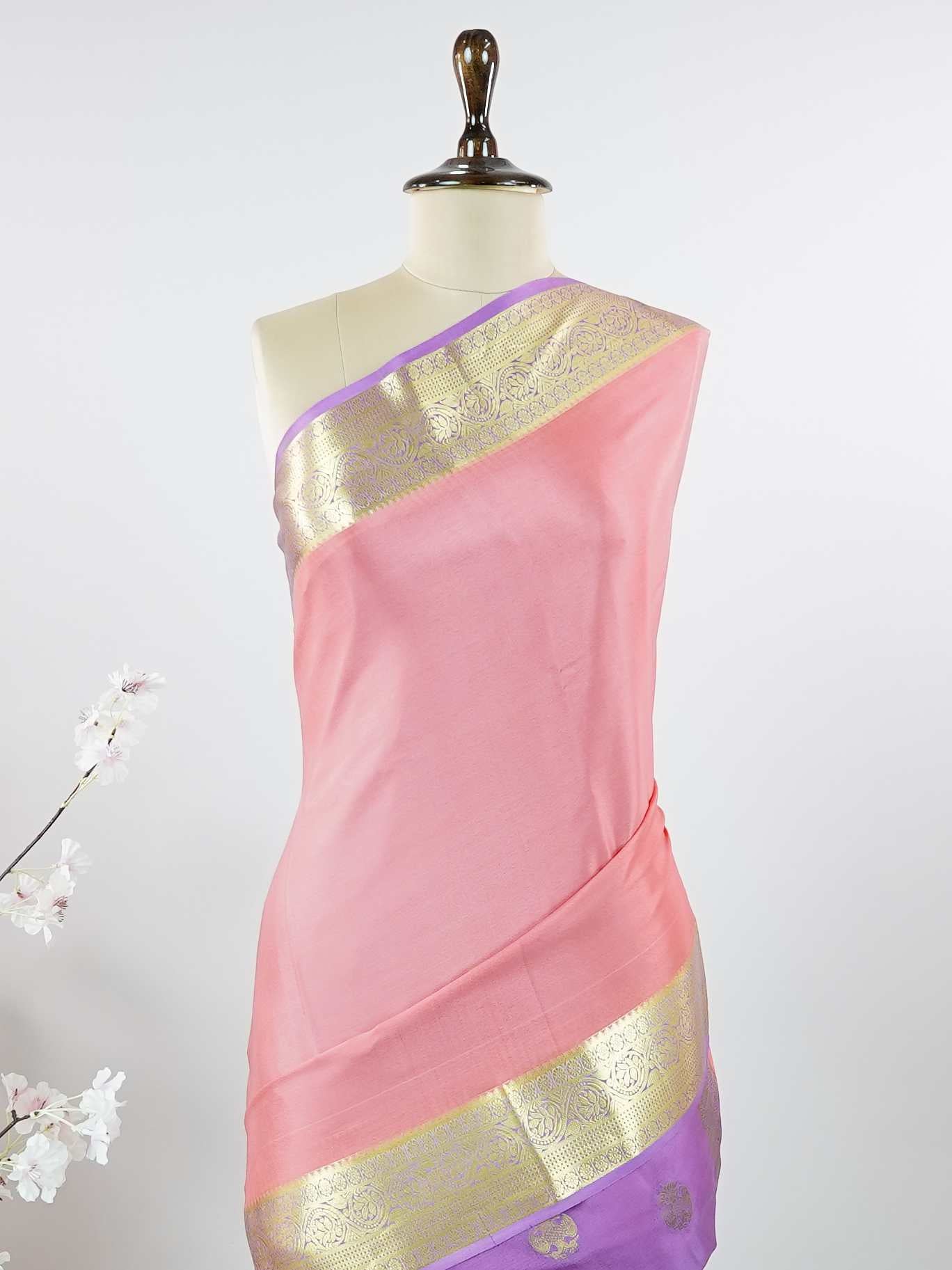 Mysore silk Gowrimanohari is a pure Mysore silk saree in the most beautiful soft rose pink — the kind of colour that feels both festive and effortlessly graceful. What makes this saree truly special is the rich purple border adorned with broad golden zari stripes that catch the light with every movement, creating a stunning contrast against the delicate pink body. The pallu carries intricate golden zari motifs woven with wonderful precision, making it a saree that looks as magnificent draped over the should