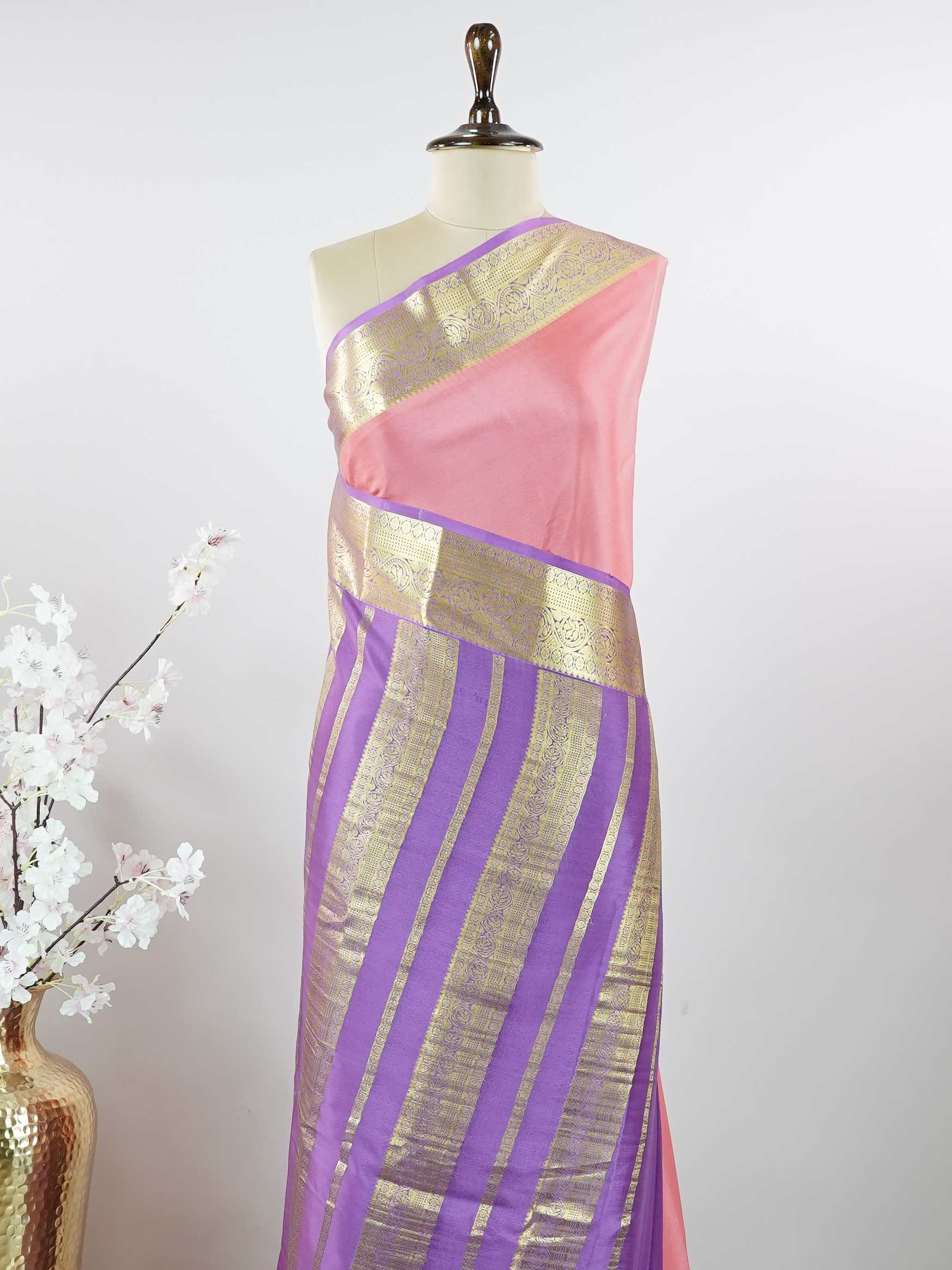 Mysore silk Gowrimanohari is a pure Mysore silk saree in the most beautiful soft rose pink — the kind of colour that feels both festive and effortlessly graceful. What makes this saree truly special is the rich purple border adorned with broad golden zari stripes that catch the light with every movement, creating a stunning contrast against the delicate pink body. The pallu carries intricate golden zari motifs woven with wonderful precision, making it a saree that looks as magnificent draped over the should