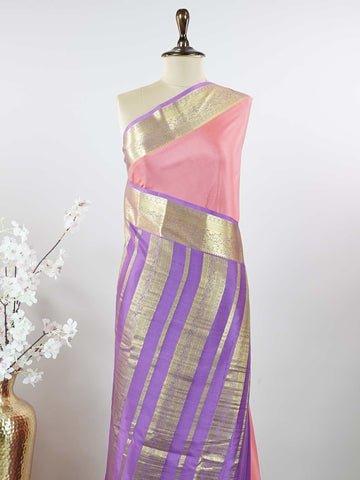Mysore silk Gowrimanohari is a pure Mysore silk saree in the most beautiful soft rose pink — the kind of colour that feels both festive and effortlessly graceful. What makes this saree truly special is the rich purple border adorned with broad golden zari stripes that catch the light with every movement, creating a stunning contrast against the delicate pink body. The pallu carries intricate golden zari motifs woven with wonderful precision, making it a saree that looks as magnificent draped over the should